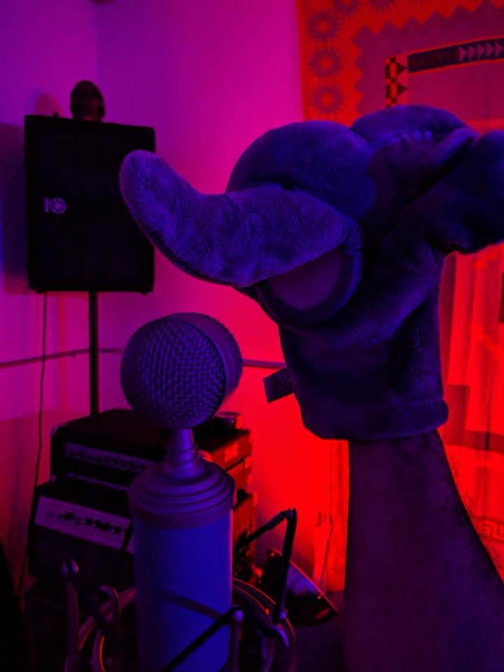 Person wearing an oversized blue mascot head at a microphone under magenta lights