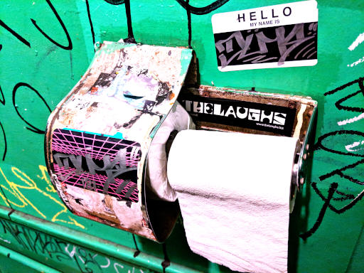 Toilet paper dispenser on a graffiti-covered wall, marked with a Laughs sticker